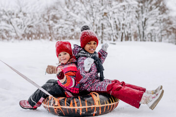 Happy Caucasian and African-American girls ride on tubing in the winter park.Beautiful trees are covered with white snow.Winter fun,active lifestyle concept.
