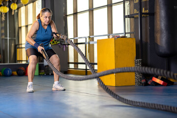 Fototapeta na wymiar Active old woman in sportswear training with battle rope in cross fit gym 