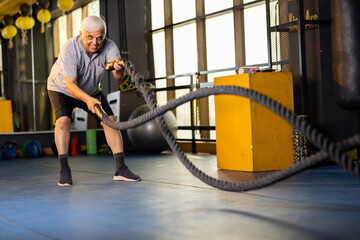 Active old man in sportswear training with battle rope in cross fit gym
