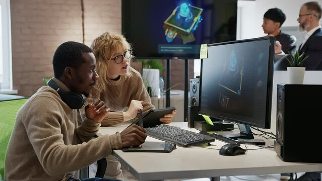 A European woman and an African American male artist discuss the 3D model in a computer game and application development studio among other team members.