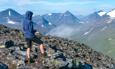 Female hiker in blue jacket overlooking vast arctic mountain landscape from the top of the mountain. Top of Naite, Sarek National Park, Sweden. Hiking in Lapland. Outdoor adventure in the arctic.