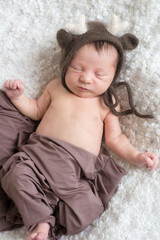 A cute little baby in a knitted hat with deer horns and a brown blanket sleeps on a white bouclé bedspread at home. Health and motherhood