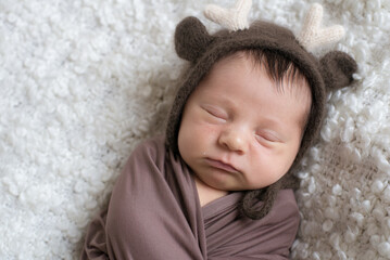 A cute little baby in a knitted hat with deer horns and a brown blanket sleeps on a white bouclé bedspread at home. Health and motherhood