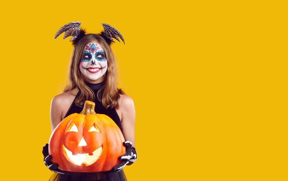 Happy child in Halloween costume on copy space background. Cheerful girl with beautiful Catrina skull makeup standing isolated on yellow background, holding jack o lantern, looking to side and smiling