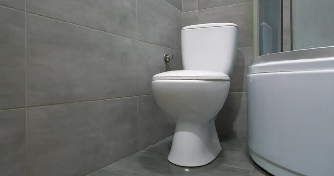Toilet room interior with white toilet bowl with closed lid, shower cabin and grey wall tiles