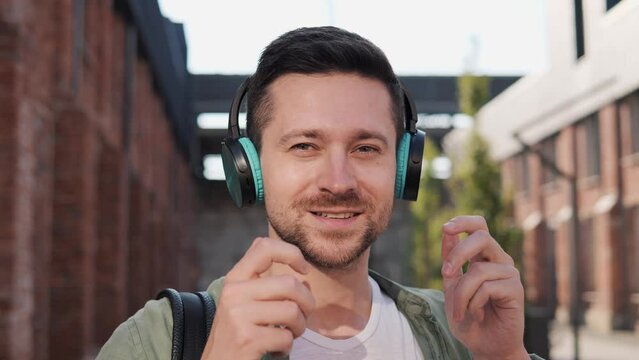 Positive man freelancer puts on wireless green headphones. Smiling man stands in urban street outdoors looks into camera smiling. Guy listening to music, enjoys good quality sound of music.