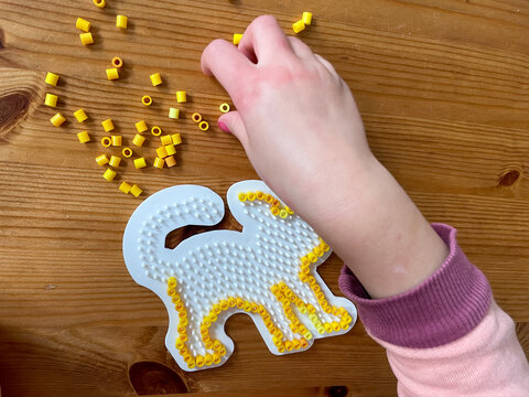 Hand eines kleinen Kinde, das gelbe Steckperlen auf eine Katzenform aufsteckt an einem Holztisch zur Besch&auml;ftigung