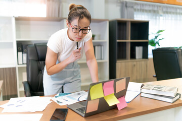 asian beautiful business woman standing thinking. serious female professional focus on thinking about information on laptop and put pen on face. occupation female worker think about document she see