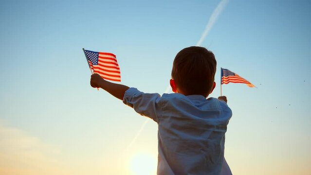 Rear view of caucasian baby boy standing and holding up his arms with two flag United States of America against blue sky. Freedom and love to country. Celebration, pride and support. Outdoor nature.