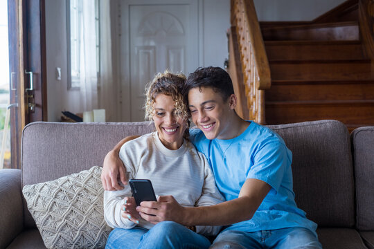 Smiling middle-aged 40s mother rest with grown-up son using smartphone together, happy young man enjoy family weekend with mom browsing wireless Internet on cellphone, have fun at home.