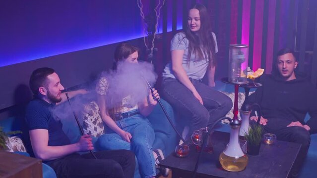 a company of young guys and girls are sitting in a hookah lounge in a relaxed atmosphere on sofas, chatting, drinking tea and smoking a hookah