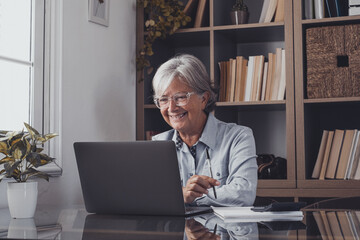 Happy old caucasian businesswoman smiling working online watching webinar podcast on laptop and learning education course conference calling make notes sit at work desk, elearning concept.