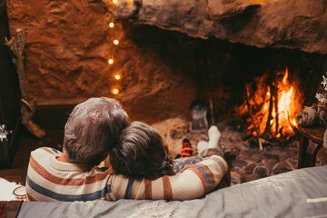 Fototapeta na wymiar Portrait of cute mature senior enjoying Christmas in front of hot fireplace at home feeling cold embracing together and holding their hands. Wearing colored holiday socks in feet..