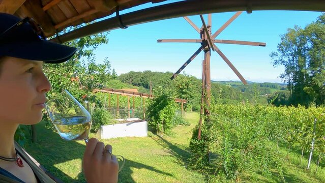 Woman holding a glass of white wine in the wine region of Gamlitz, Southern Styria (Steiermark), Austria, Europe. Scenic view on a vineyard and a local bird scarer windmill called klopotec (klapotetz)