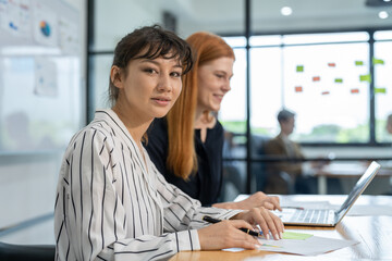 In the office, two businesswomen are using laptops to discuss marketing report documents and exchange ideas. Plan strategies for business team meetings.