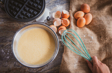 Making waffles homemade with waffle iron, fresh waffle in bowl, eggs and hands holding light blue whisk.