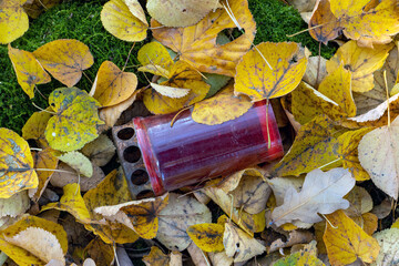 A fallen memorial candlestick lies in grass covered with fallen leaves at autumn cemetery
