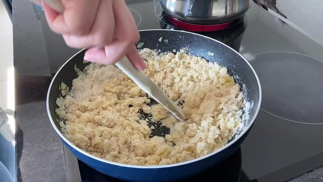SLOW MOTION SHOT - Preparing farofa (Brazilian toasted cassava flour) toasting in a pan on the electric stove. Adding the cassava flour and stirring continuously with a wooden spoon.
