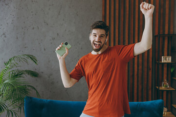 Young joyful man in red t-shirt listen music in earphones use mobile cell phone dancing near blue sofa stay at home flat rest relax spend free spare time in living room indoor People lounge concept.