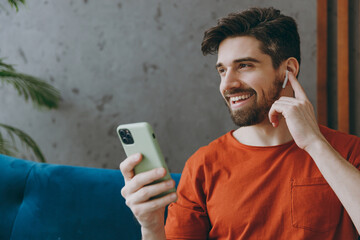 Young man wears red t-shirt listen music in earphones use mobile cell phone sit on blue sofa stay...