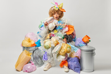 Plastic pollution concept. Scared emotional curly woman overloaded with plastic garbage shocked to collect so much wastes finds out about big environmental disaster isolated over grey background