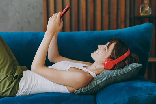 Side view young smiling happy woman wear white tank shirt headphones listen music use mobile cell phone lay down on blue sofa stay at home flat rest relax spend free spare time in living room indoor.