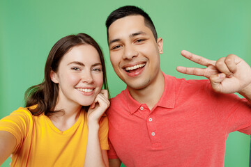 Close up young happy couple two friends family man woman wear basic t-shirts together do selfie shot pov on mobile cell phone show v-sign gesture isolated on pastel plain light green color background.