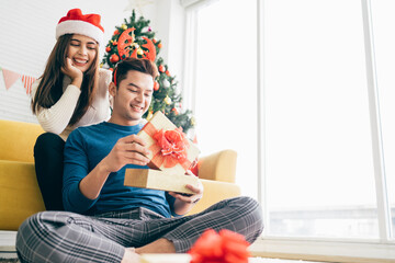 Young beautiful happy Asian woman wearing Santa Claus hat surprises her boyfriend with a Christmas gift at home with Christmas tree in the background. Image with copy space.