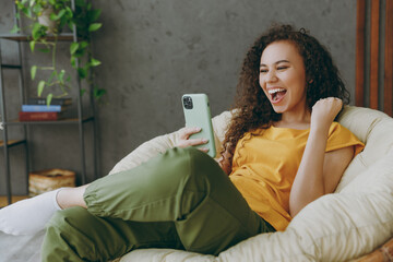 Young fun woman of African American ethnicity wear t-shirt use mobile cell phone do winner gesture sits in armchair stay at home flat rest relax spend free spare time in living room indoors grey wall.