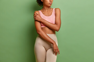 Cropped shot of unrecognizable woman keeps hand on shoulder dressed in cropped top and leggings demonstrates perfect figure well cared skin isolated over vivid green background. Body care concept
