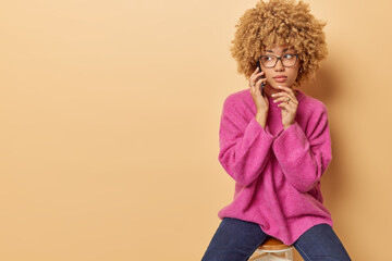 Indoor shot of beautiful young woman with curly hair holds hand on chin has telephone talk looks aside with serious expression wears pink jumper and jeans poses on chair against beige background