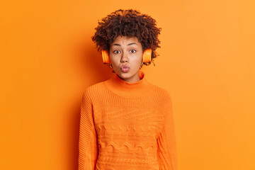 Fototapeta na wymiar Horizontal shot of lovely curly haired woman keeps lips folded has romatic mood listens music via stereo headphones enjoys favorite song dressed in casual jumper isolated over orange background