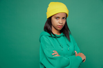 Side view sad little kid teen girl of African American ethnicity 13-14 years old wear casual hoody hat hold hands crossed folded look camera isolated on plain dark green background Childhood concept.