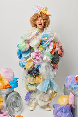 Vertical shot of female volunteer collects plastic garbage cleans up litter makes costume from wastes laughs joyfully stands in full length against grey background. Environmental conservation