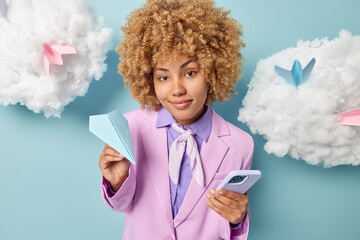 Gorgeous curly haired woman entrepreneur holds handmade paper plane modern cellular checks notification dressed formally gets ready for formal meeting arranges meeting isolated over blue background.