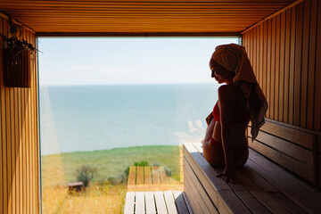 woman in a red swimsuit sits on a bench in the sauna with the panoramic window. 