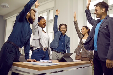 Team of happy diverse business people celebrating success and having fun in a work meeting. Group of cheerful, confident men and women standing around an office table, raising hands up and smiling