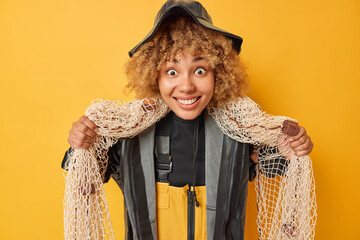 Positive curly fisherwoman focused at camera with happy expression carries fishing net around neck...