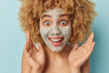 Portrait of surprised cheerful woman with curly blonde hair keeps palms raised stares shocked at camera smiles broadly undergoes beauty procedures applies clay mask isolated over blue background