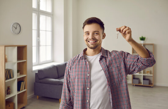 Indoor shot of happy home owner with key to his new house. Cheerful handsome single young Caucasian man in casual shirt standing in living room, showing keys and smiling. Real estate, purchase concept