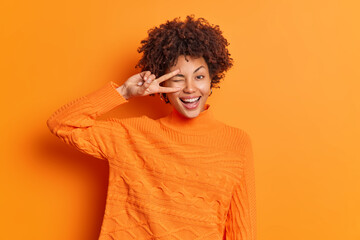 Indoor shot of happy curly haired woman makes peace gesture over eye shows v sign smiles toothily dressed in casual jumper isolated over vivid orange background. People body language and fun concept