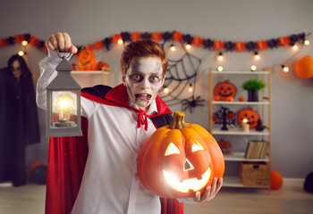 Portrait of child in spooky vampire costume. Kid dressed as Count Dracula holding lamp and jack-o-lantern. Teen boy in cape with crazy eyes and fake blood makeup looking for candy on Halloween night