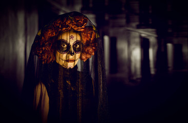 Portrait of mystical Calavera Catrina with sugar skull makeup on dark gloomy background. Close up of woman in Day of the Dead costume wearing floral wreath and black veil looking directly into camera.