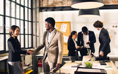 Selective focus African handsome businessman and Asian beautiful businesswoman colleague wearing formal clothes, greeting, talking each other, smiling with happiness while standing in indoor office.