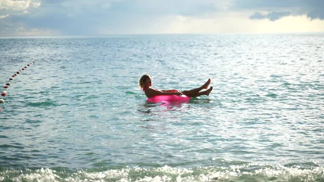Funny woman with a laptop at the sea in an inflatable circle. View from the shore of a blonde woman floating on an inflatable big pink donut with a laptop in the sea at sunset