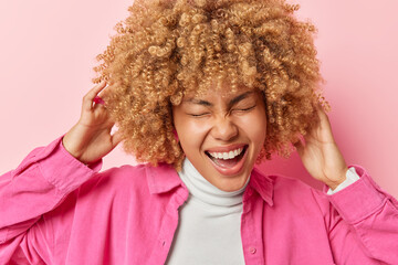 Portrait of cheerful woman laughs happily keeps hands on curly hair keeps eyes closed from joy dressed in casual clothes poses against pink background. People positive emotions and feelings concept