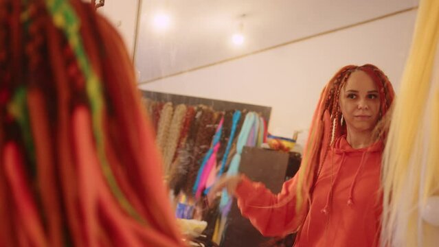 Young beautiful woman looking in mirror, considering dreadlocks, touching hairstyle, preening.