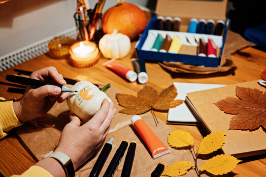 Autumn pumpkins craft. DIY Fall Crafts for Adults. Female hands draw patterns on white pumpkin against autumn decor background on the table at home