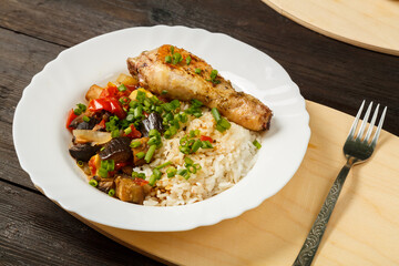 Rice with vegetables and chicken leg on a handmade wooden board next to a fork on a wooden table.