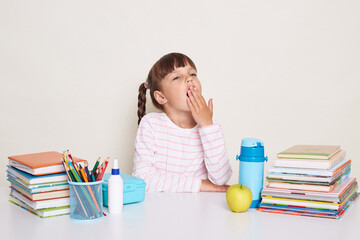 Photo of sleepy tired little schoolgirl with pigtails wearing striped shirt sitting at the desk surrounded school supplies, covering mouth with palm, yawning, being sleepless.